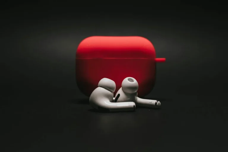 A red charging case open beside a pair of white wireless earbuds on a black background