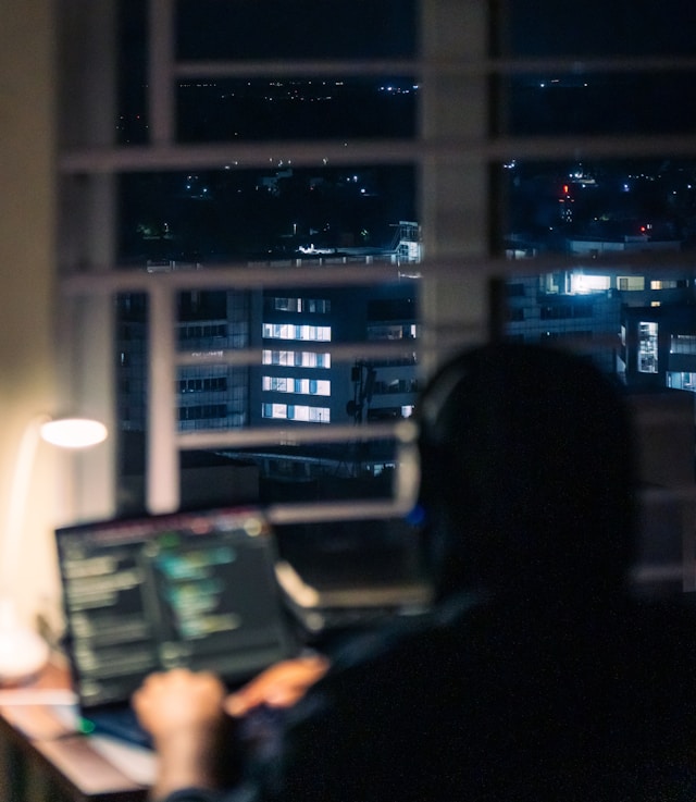 A person silhouetted against a window at night in a dark room, with city lights visible outside