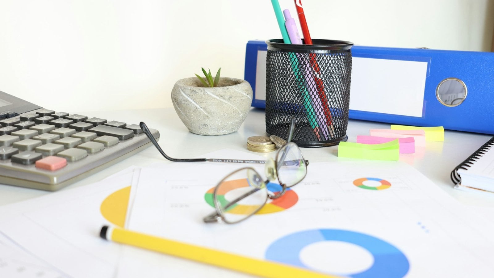 A desk flat lay with a calculator, ring binders, a pen pot, glasses and sticky notes