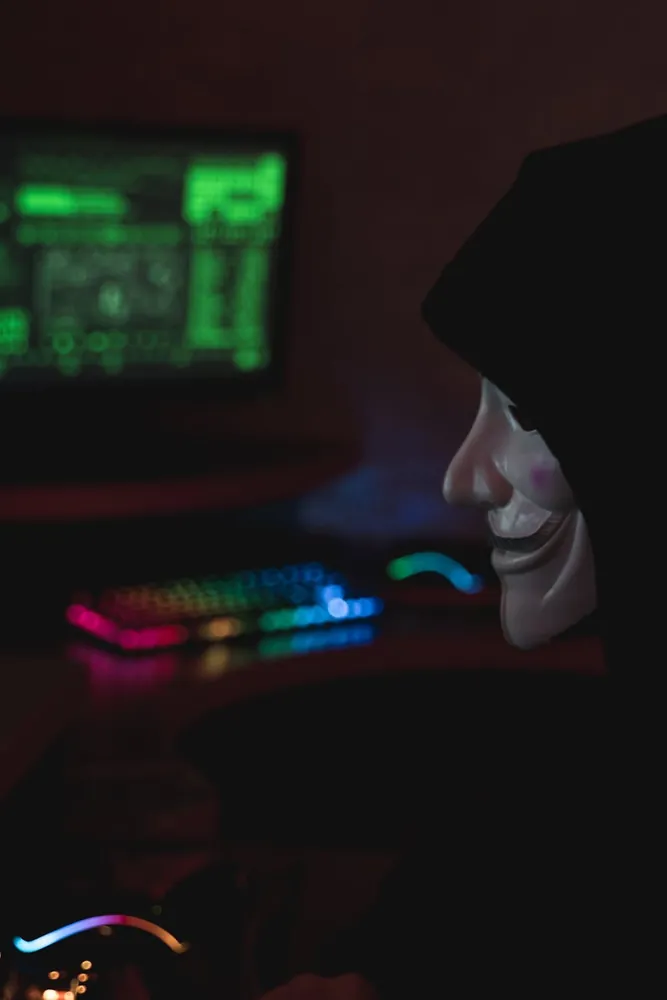 A figure in a pale mask sitting in the dark behind a backlit keyboard