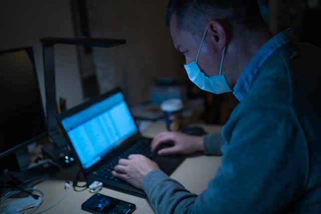 A healthcare worker in a face mask working at two computer screens in a dim room