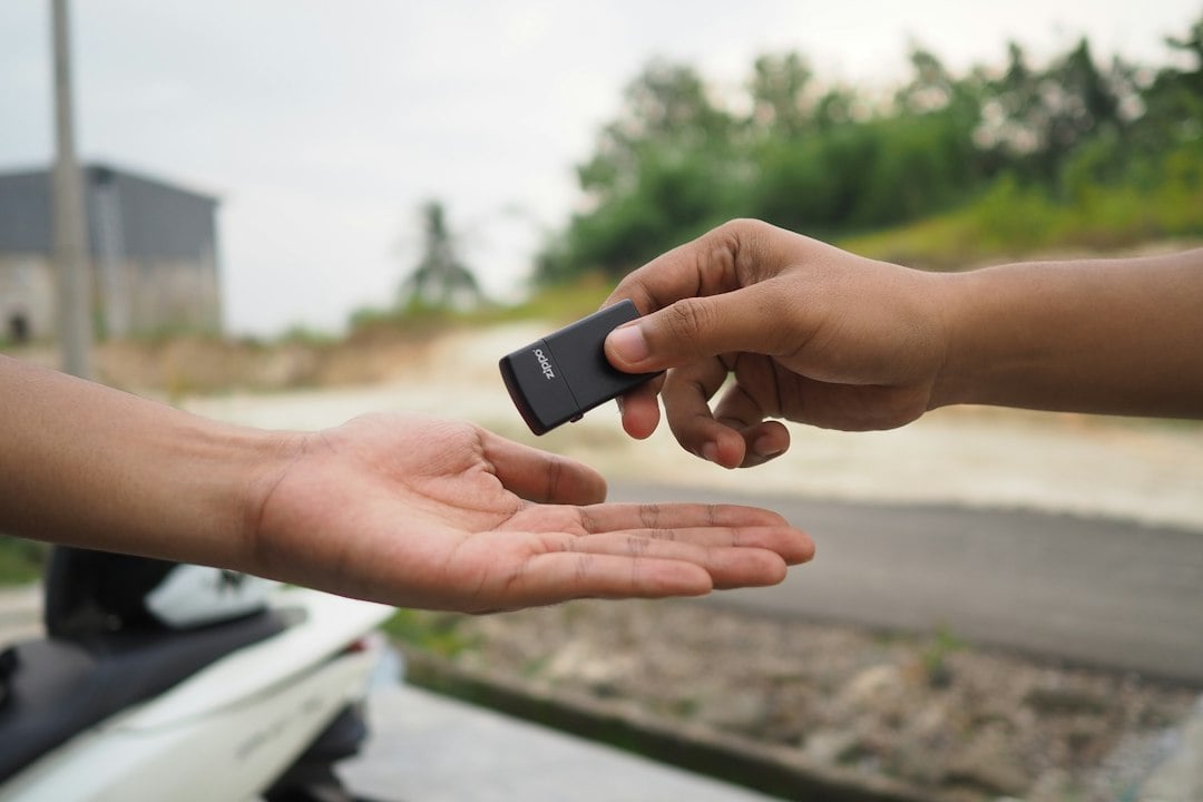 A hand passes a small black key fob into another person's open palm outdoors