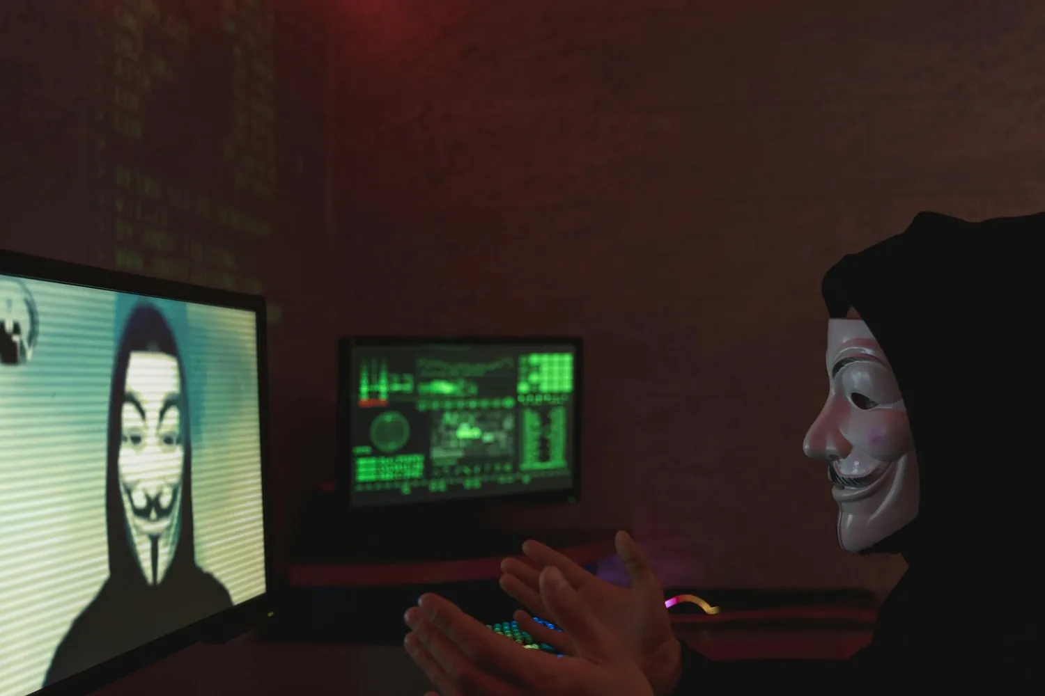 A person wearing a Guy Fawkes mask sitting in a dark room in front of glowing monitors