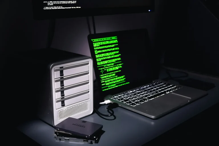 A laptop and an external drive on a desk in a dark room, green code filling the laptop screen