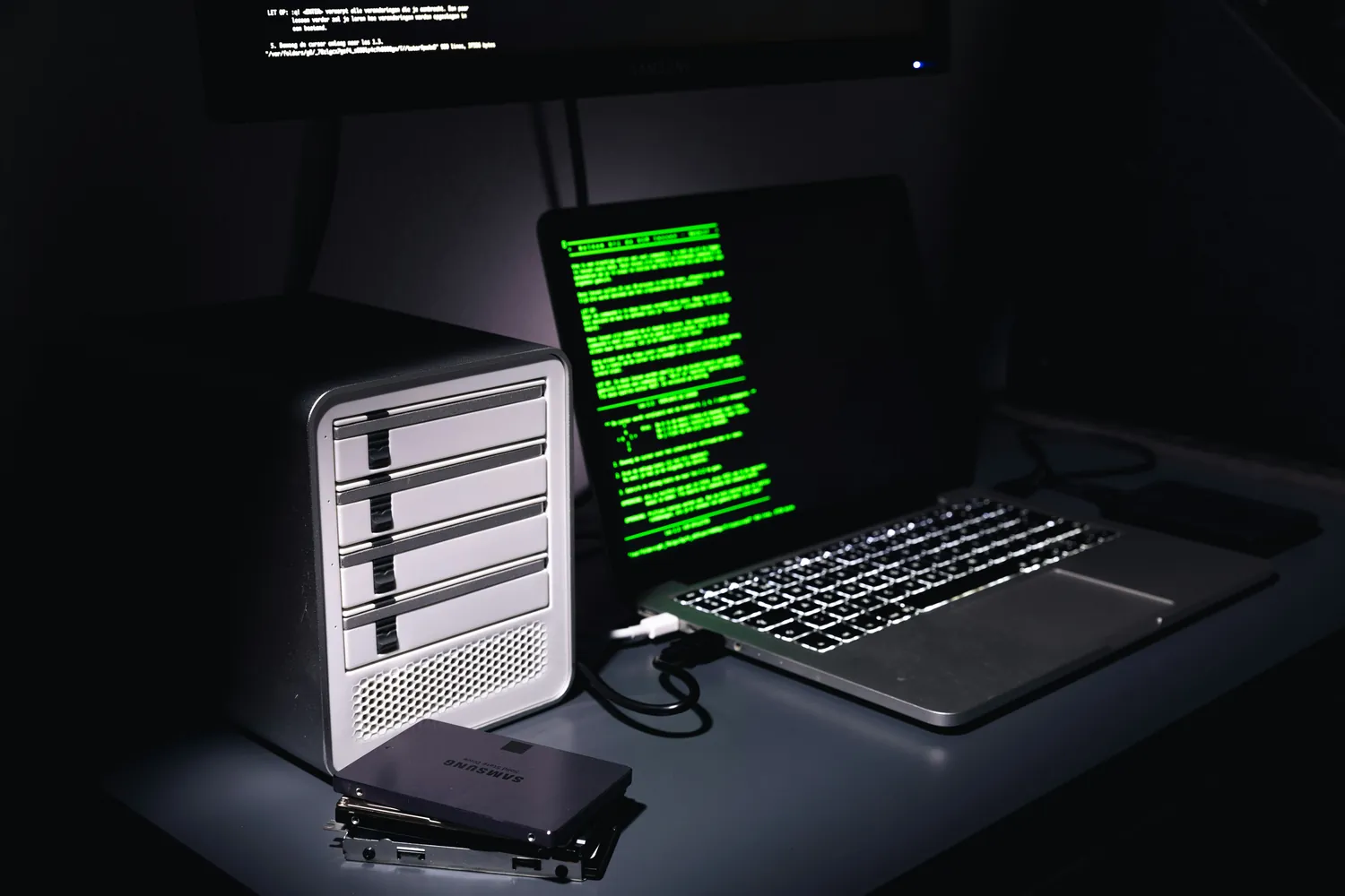 A laptop and an external drive on a desk in a dark room, green code filling the laptop screen