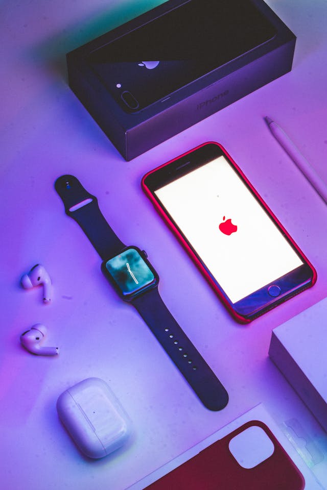 An Apple Watch, wireless earbuds and a phone on a purple surface lit in pink light
