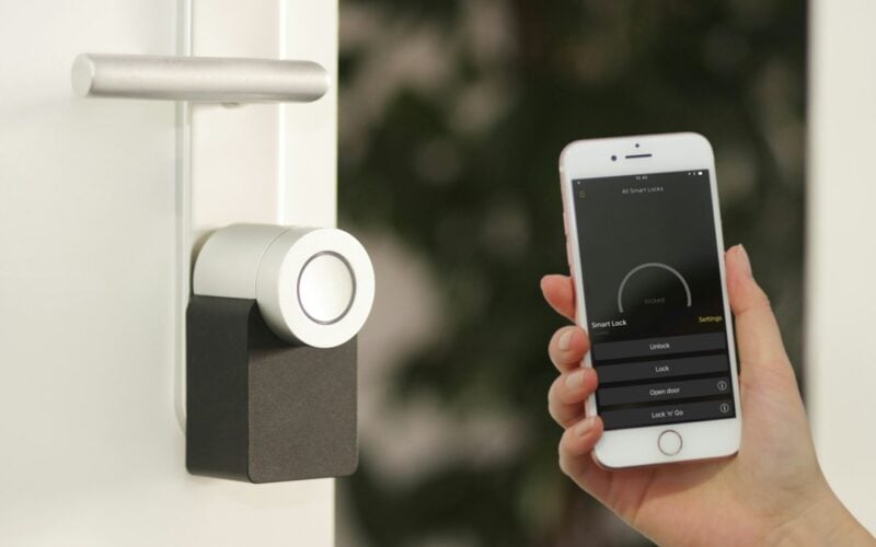 A hand holds a smartphone running a lock app beside a smart deadbolt mounted on a white front door.