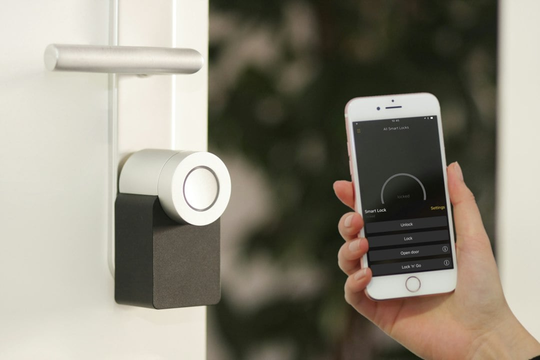 A hand holds a smartphone running a lock app beside a smart deadbolt mounted on a white front door.