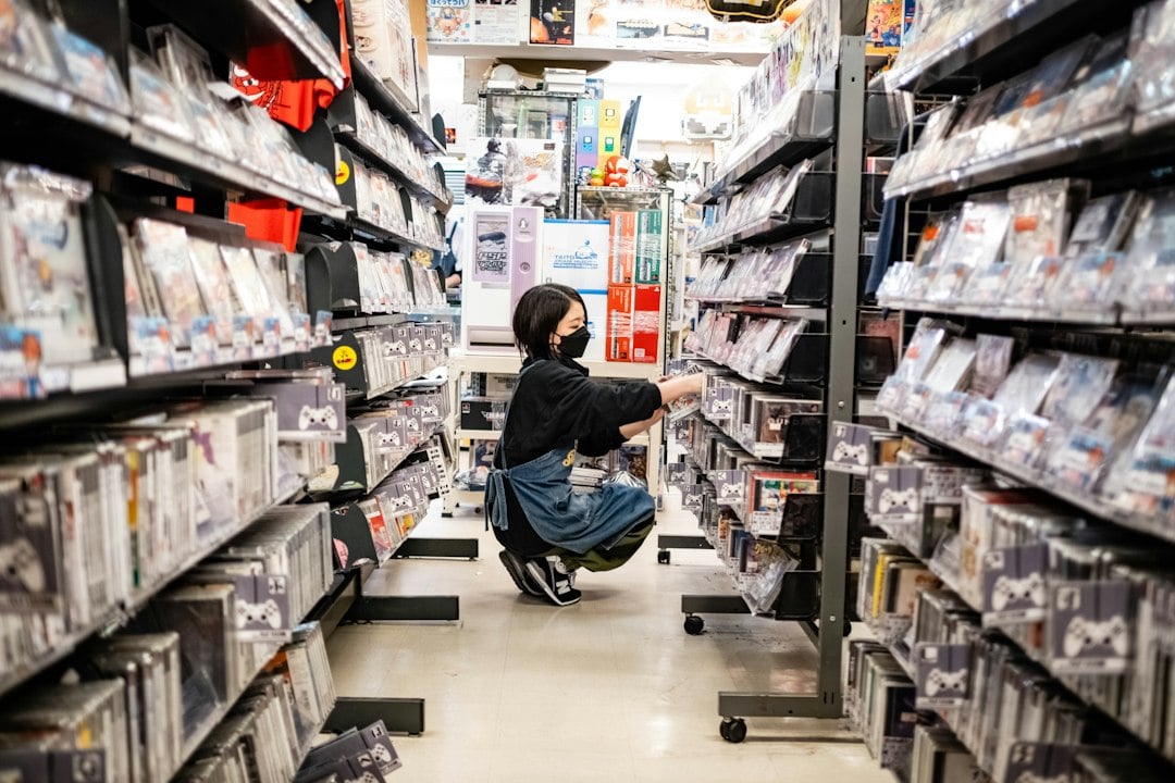 A customer crouches to inspect boxed products on densely stocked shelves in a narrow store aisle