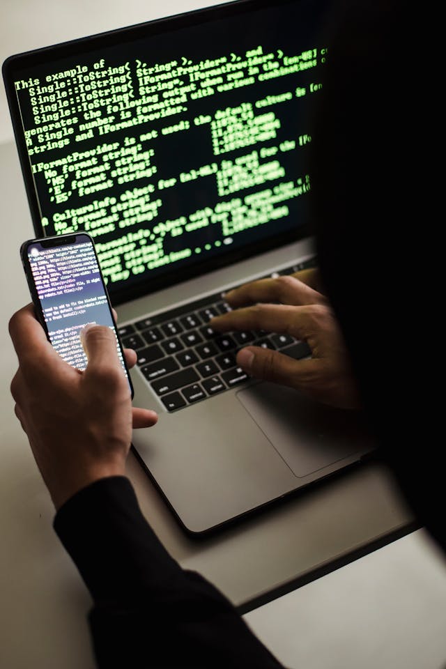 A hand holding a phone in front of a laptop keyboard, with green code filling the laptop screen