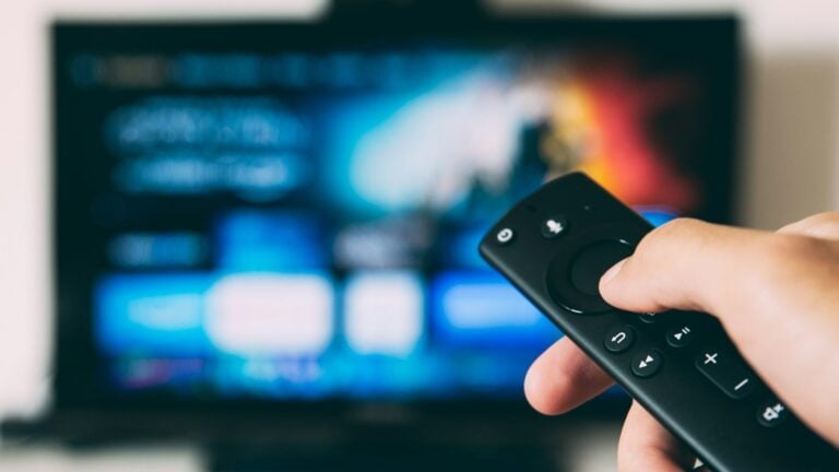 A hand holding a television remote pointed at a screen showing a blurred streaming menu
