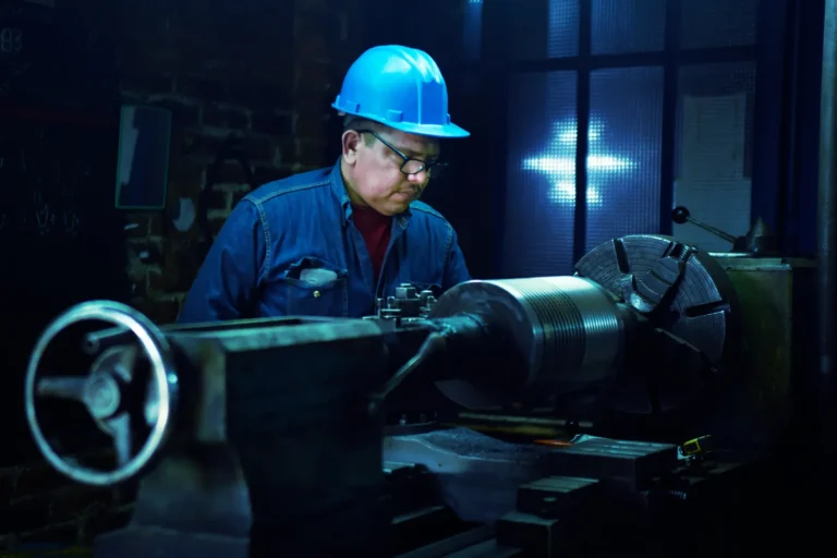 A worker in a blue hard hat and safety glasses operating a lathe in a dimly lit workshop