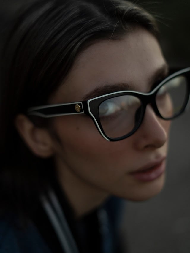 A close-up of a woman wearing dark-framed smart glasses with a small camera in the corner of the frame