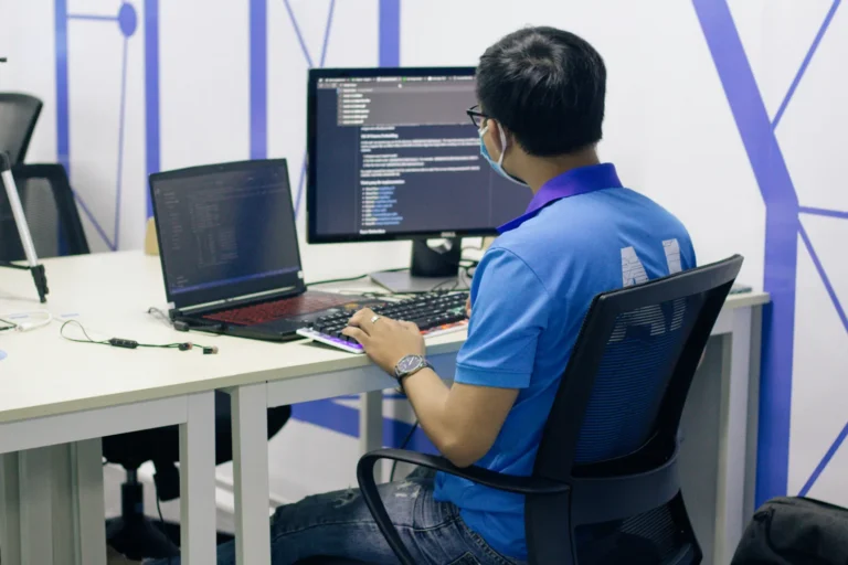 A person in a blue shirt working at a desk with a laptop and a monitor showing code