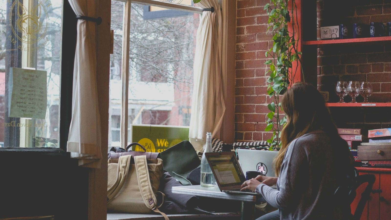 A woman seen from behind working on a laptop at a bench beside a cafe window